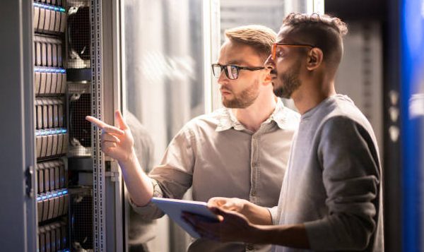 Team of young multi-ethnic specialists in glasses standing at server and using tablet while managing network server together in data center