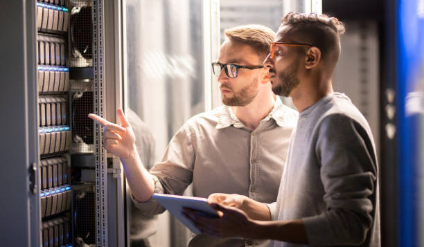 Team of young multi-ethnic specialists in glasses standing at server and using tablet while managing network server together in data center